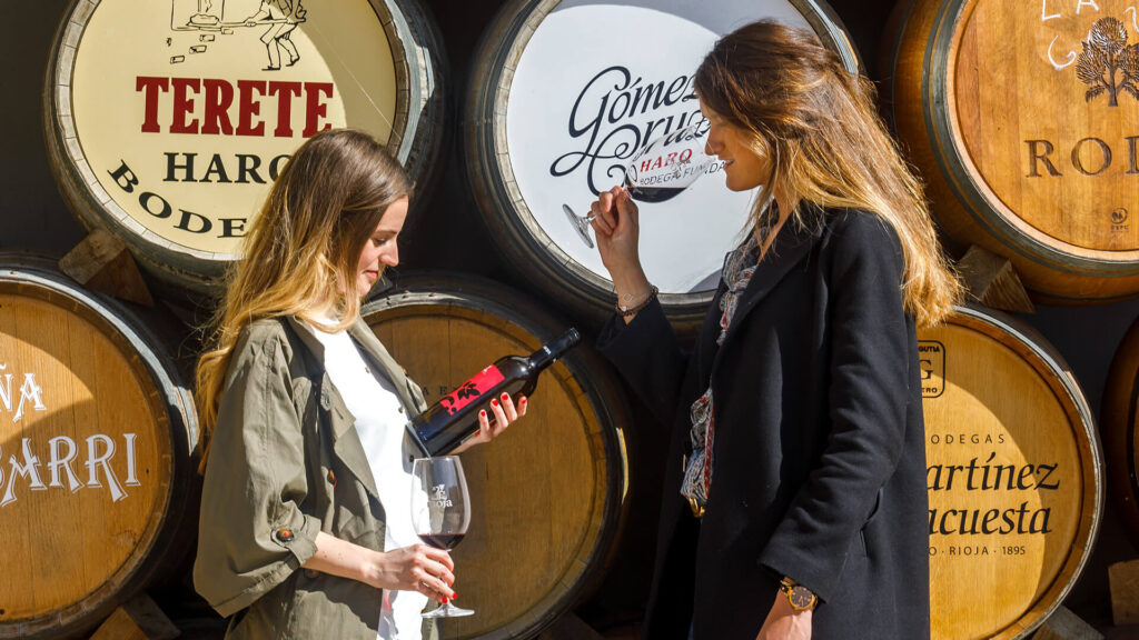two women in front of wine barrels looking and drinking a bottle of Rioja red wine