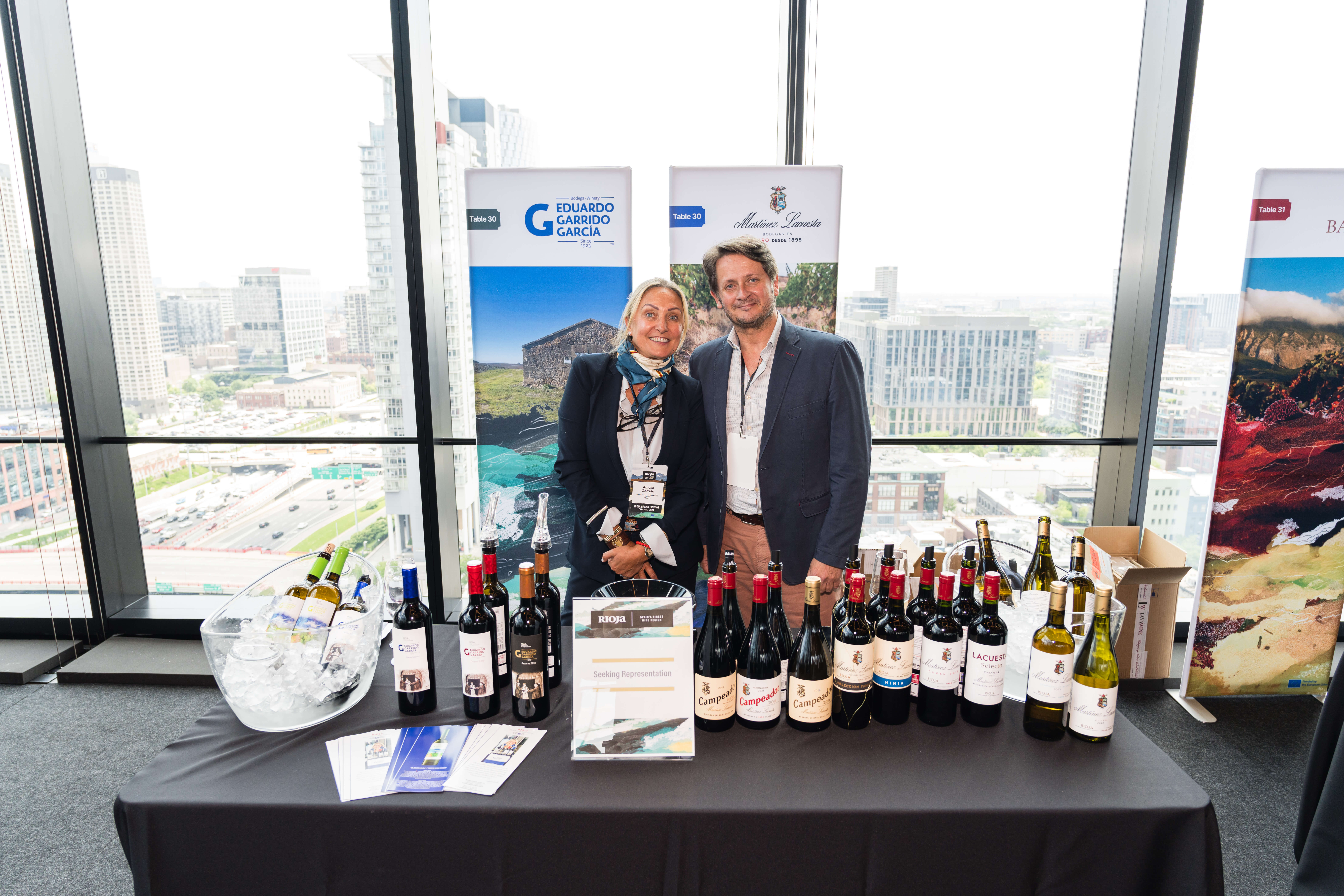 two wine professionals stand behind a table display smiling