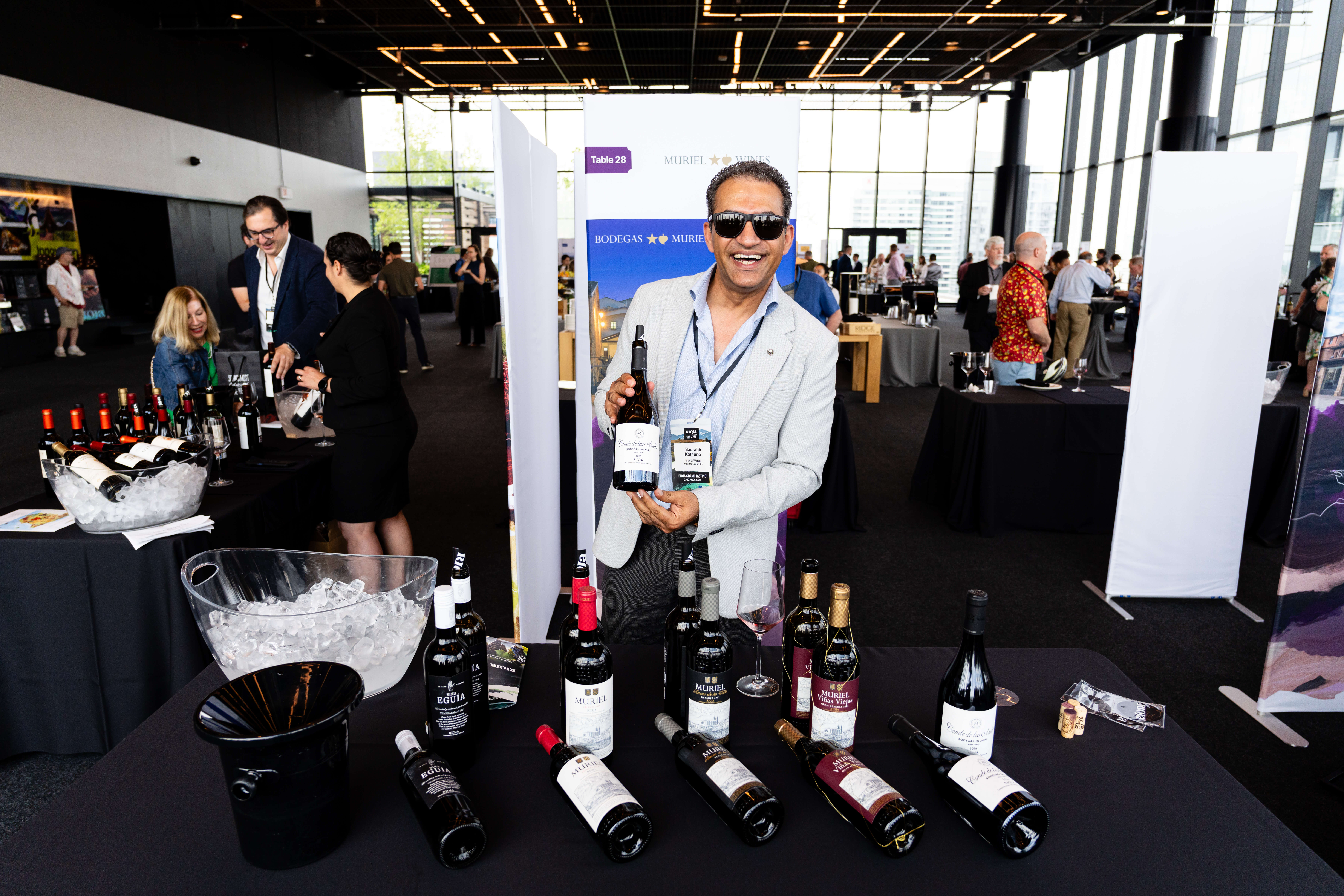 A man is smiling in sunglasses as he shows a bottle of red wine while stands at a table display